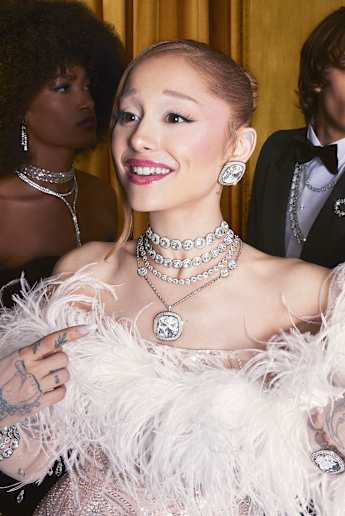 Ariana Grande wearing a feathered dress with stacked clear crystal Swarovski jewelry, surrounded by four people.