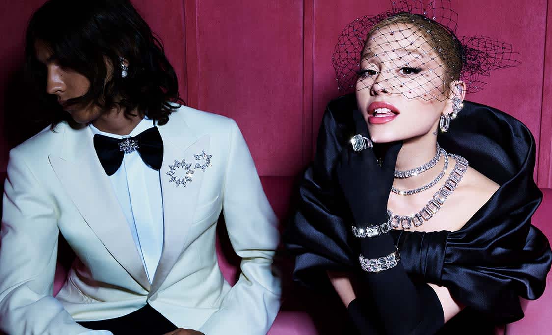 Ariana Grande w czarnym stroju i biżuterii Swarovski siedząca obok mężczyzny w smokingu