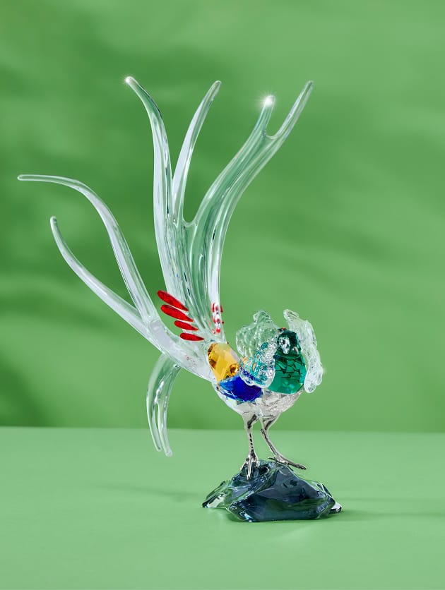 Pestrobarevná figurka z Křišťálu Swarovski stojícího ptáka s dlouhými, rozevlátými ocasními pery z čirého křišťálu, se žlutou hlavou a červenými akcenty na tmavém podstavci oproti zelenému pozadí.