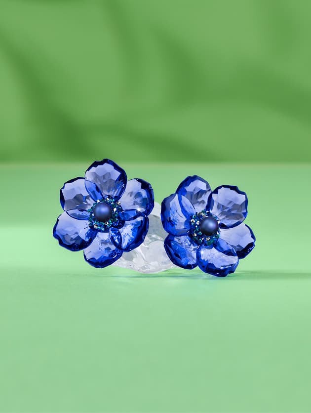 Dva malé květy z modrého Křišťálu Swarovski s vrstvenými okvětními lístky a tmavými středy na světle zeleném povrchu oproti měkce zelenému pozadí.