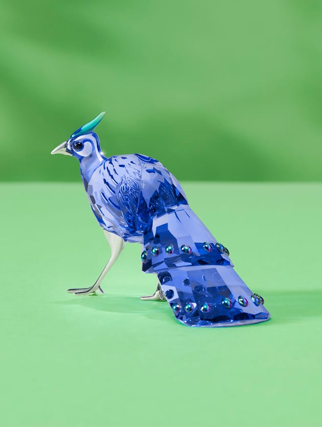 Sytě modrá figurka stojícího ptáka z Křišťálu Swarovski s hladkým tělem a elegantními ocasními pery na zeleném pozadí.